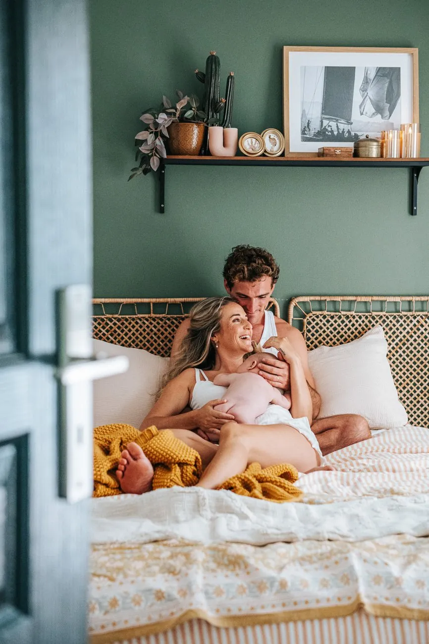 Séance bébé à domicile vue par la porte : parents riant sur leur lit avec leur bébé dans une chambre verte, dans le Var