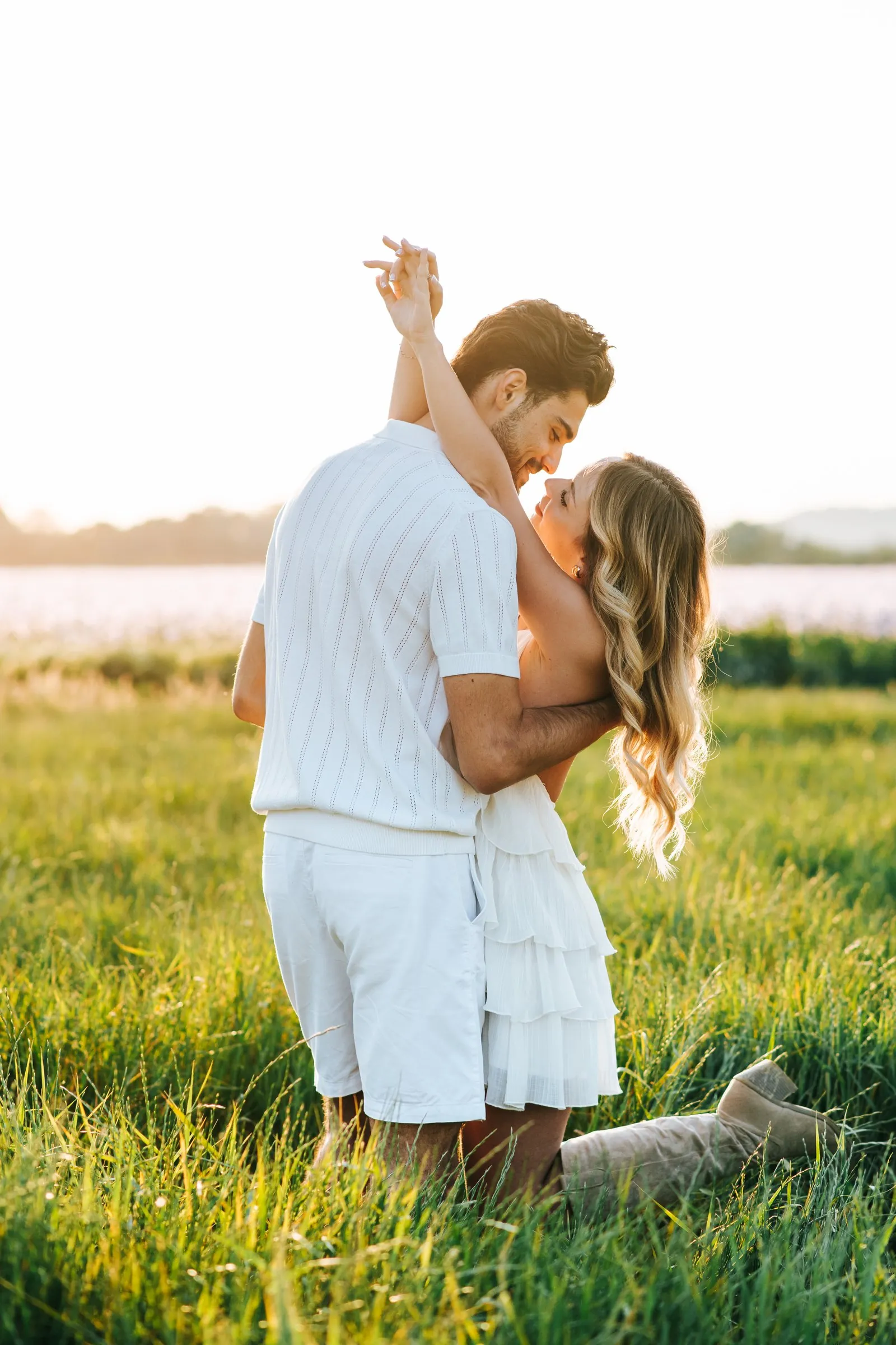 Couple session in a field at sunset, both dressed in white, her arms raised and eyes locked together