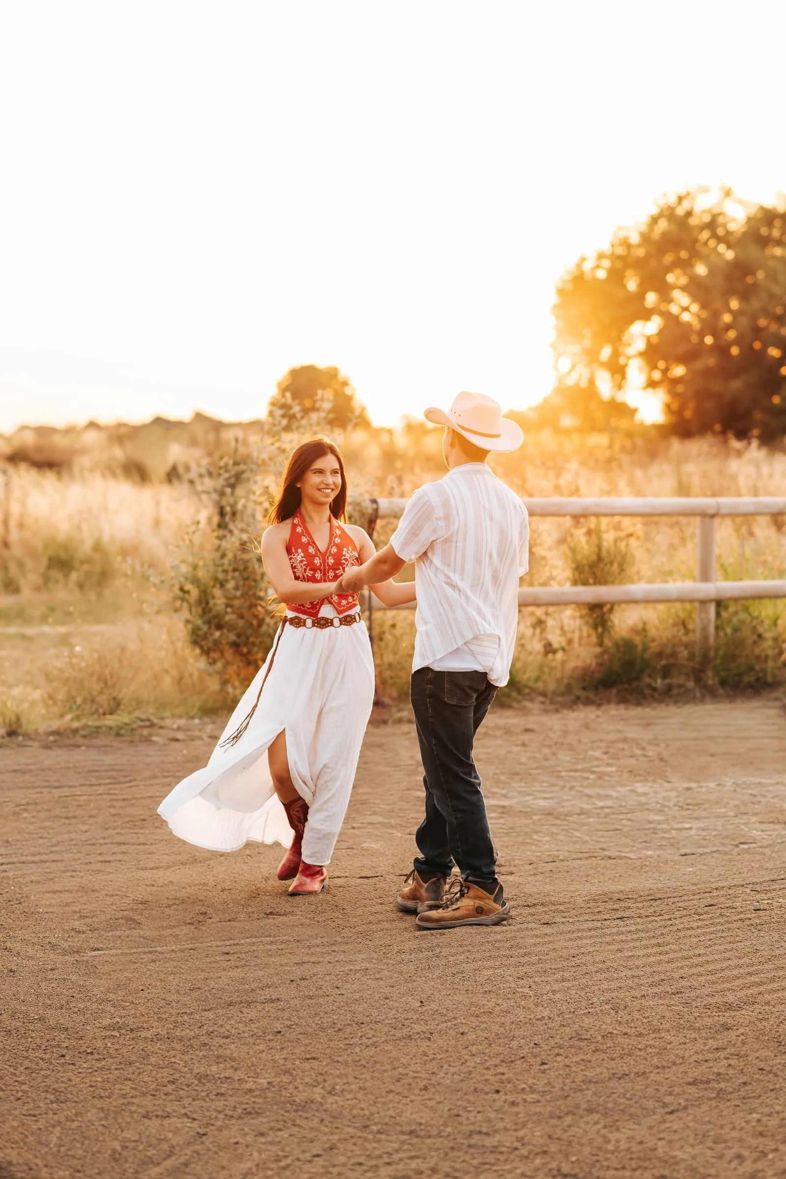 Séance couple au coucher du soleil : ils dansent main dans la main, robe blanche et chapeau de cowboy