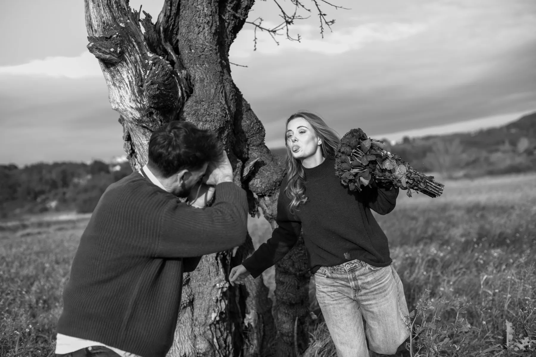 Séance couple pleine d'humour près d'un vieil arbre : elle tire la langue, bouquet de roses à l'épaule, noir et blanc