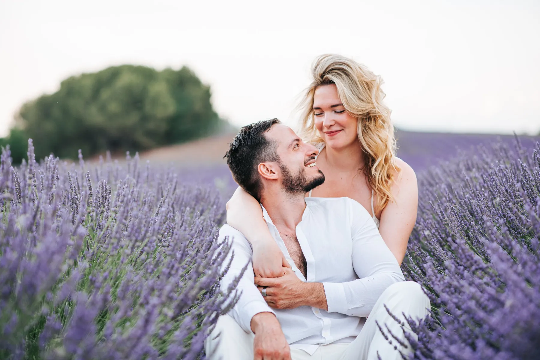 Séance couple dans les champs de lavande : elle l'enlace par derrière et ils se sourient, tenues blanches