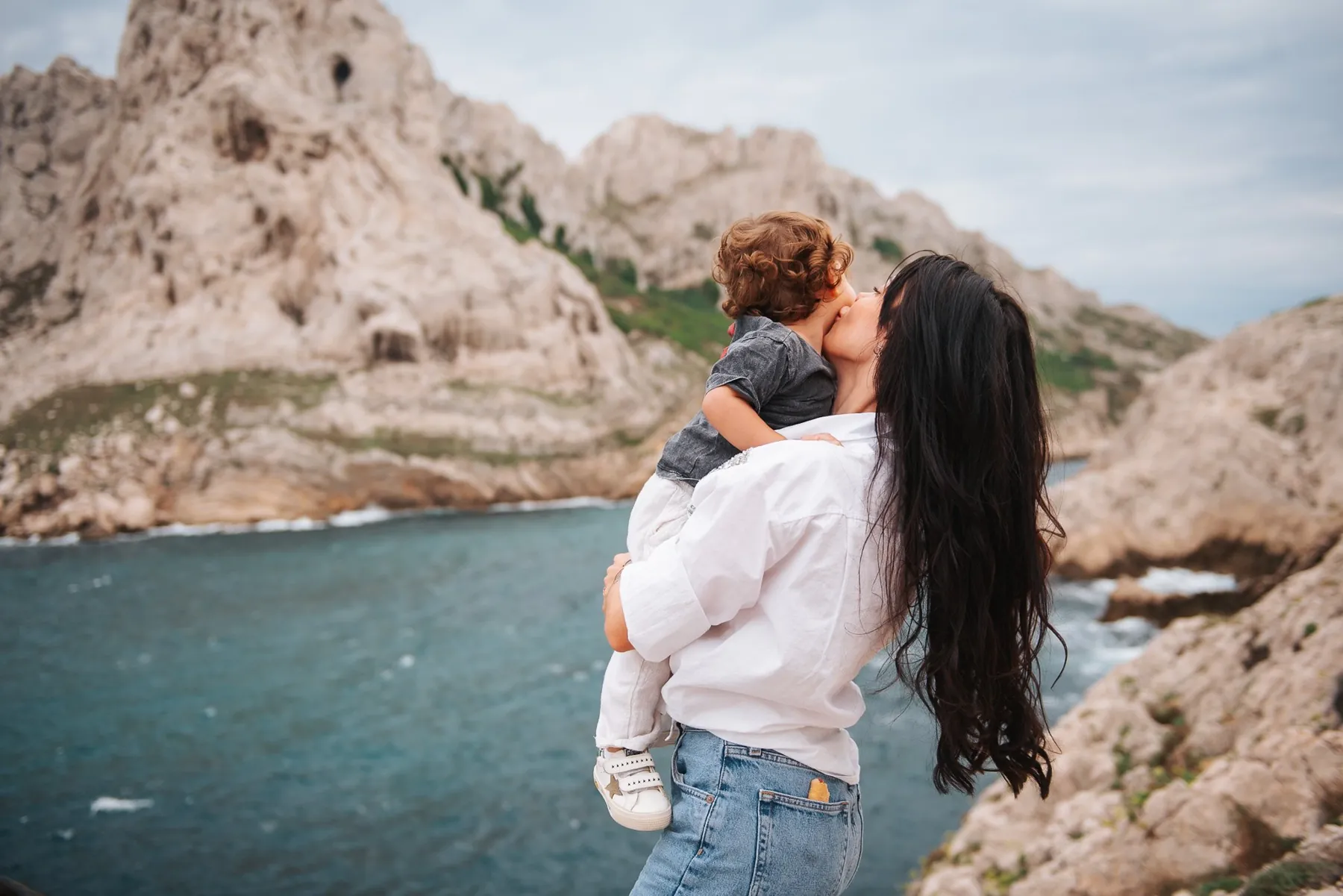 Family session in the calanques: a mum seen from behind kissing her curly-haired little boy above the turquoise sea