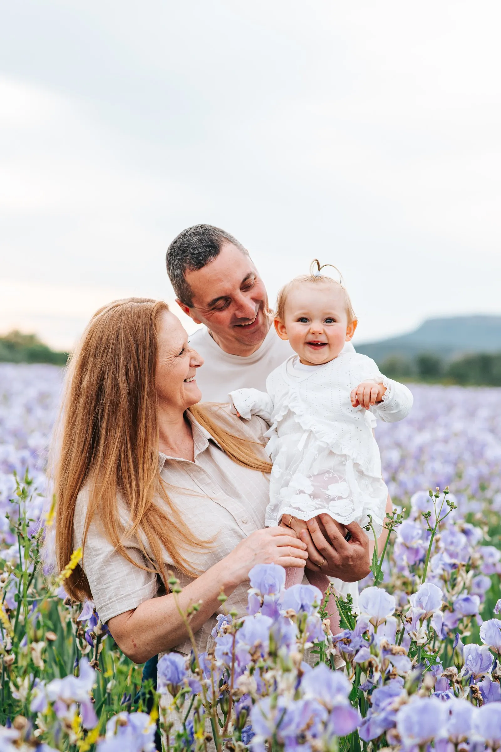 Family session in an iris field: a beaming baby in a white dress held by her laughing parents near Aix-en-Provence