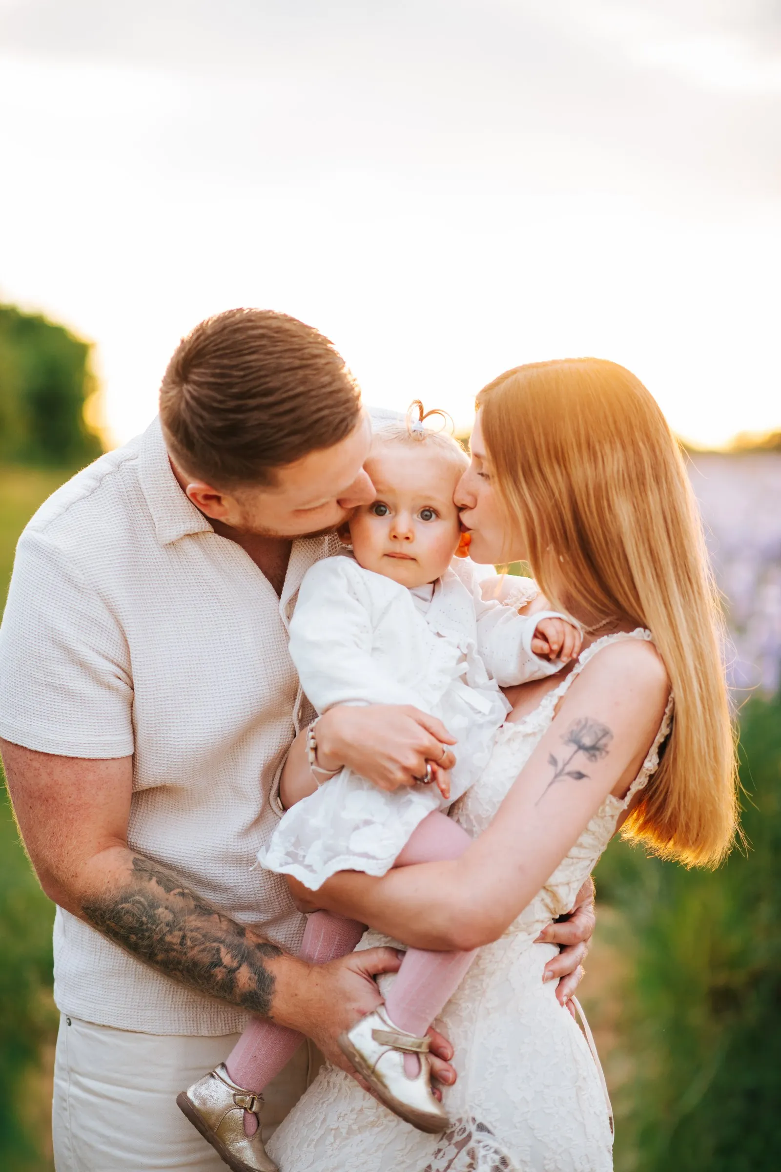 Family session in neutral tones: parents dressed in white kissing their baby's cheeks at sunset