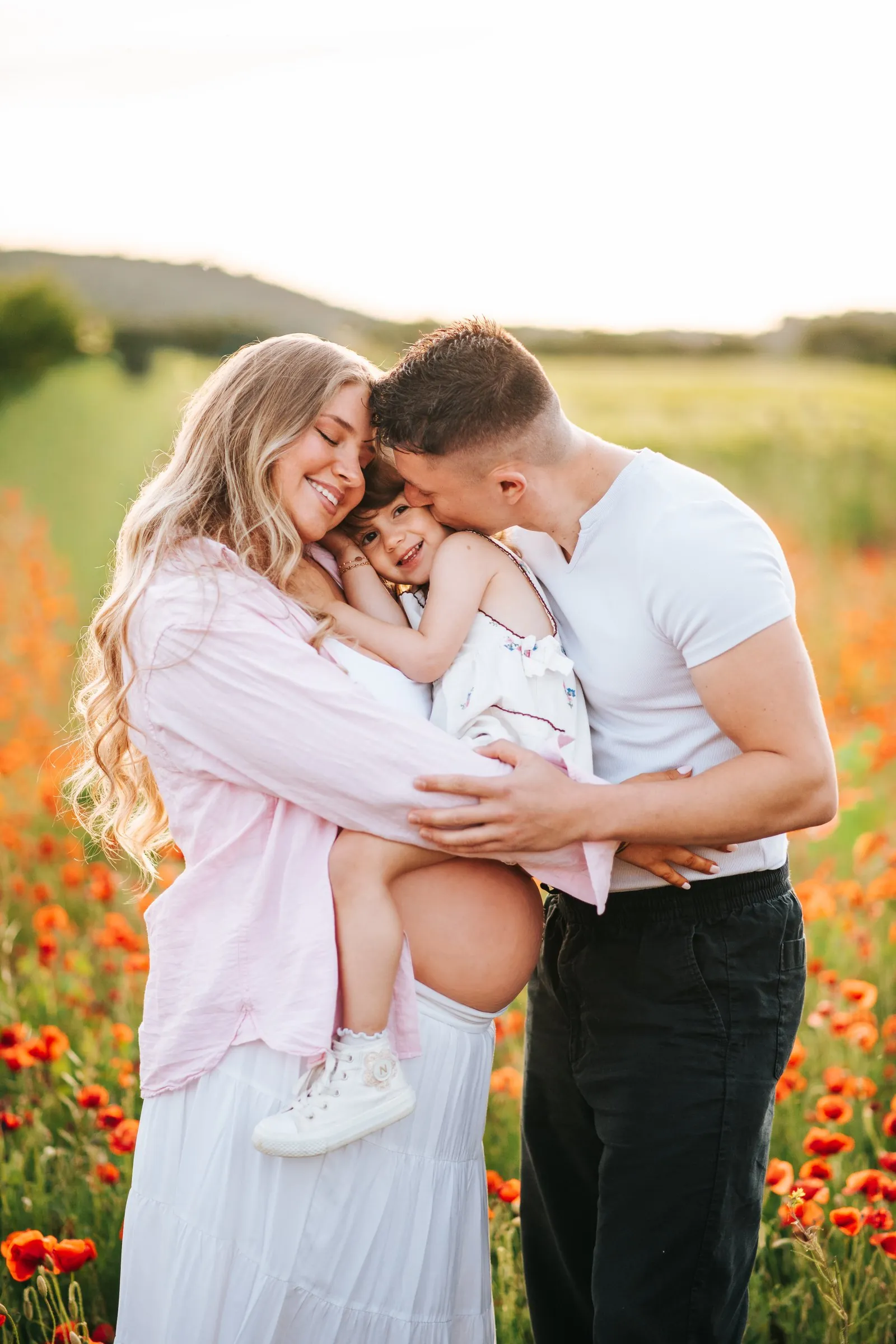 Family maternity session in a poppy field: the mum-to-be, dad and little girl cuddled up tenderly