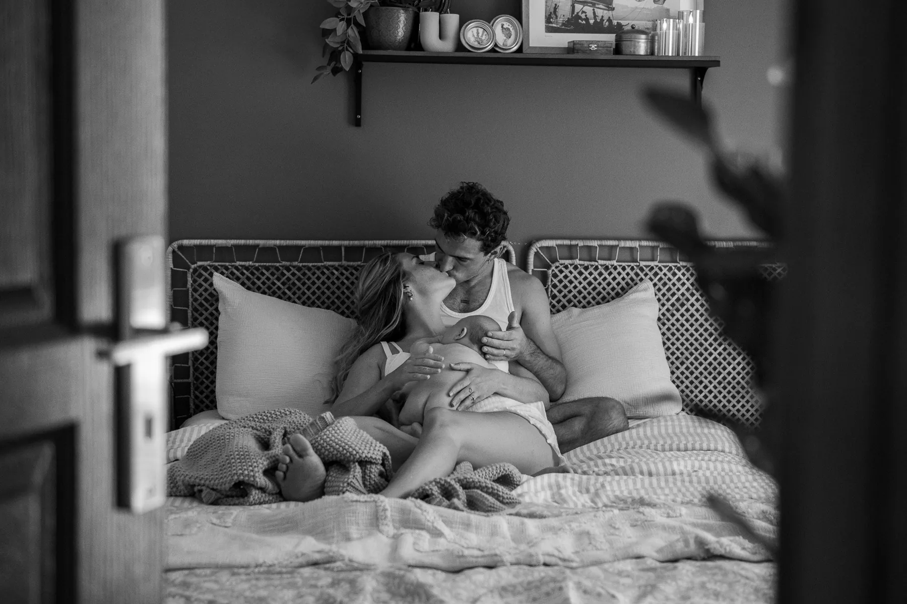 Parents qui s'embrassent sur leur lit, leur nouveau-né blotti contre maman, vus par la porte, en noir et blanc