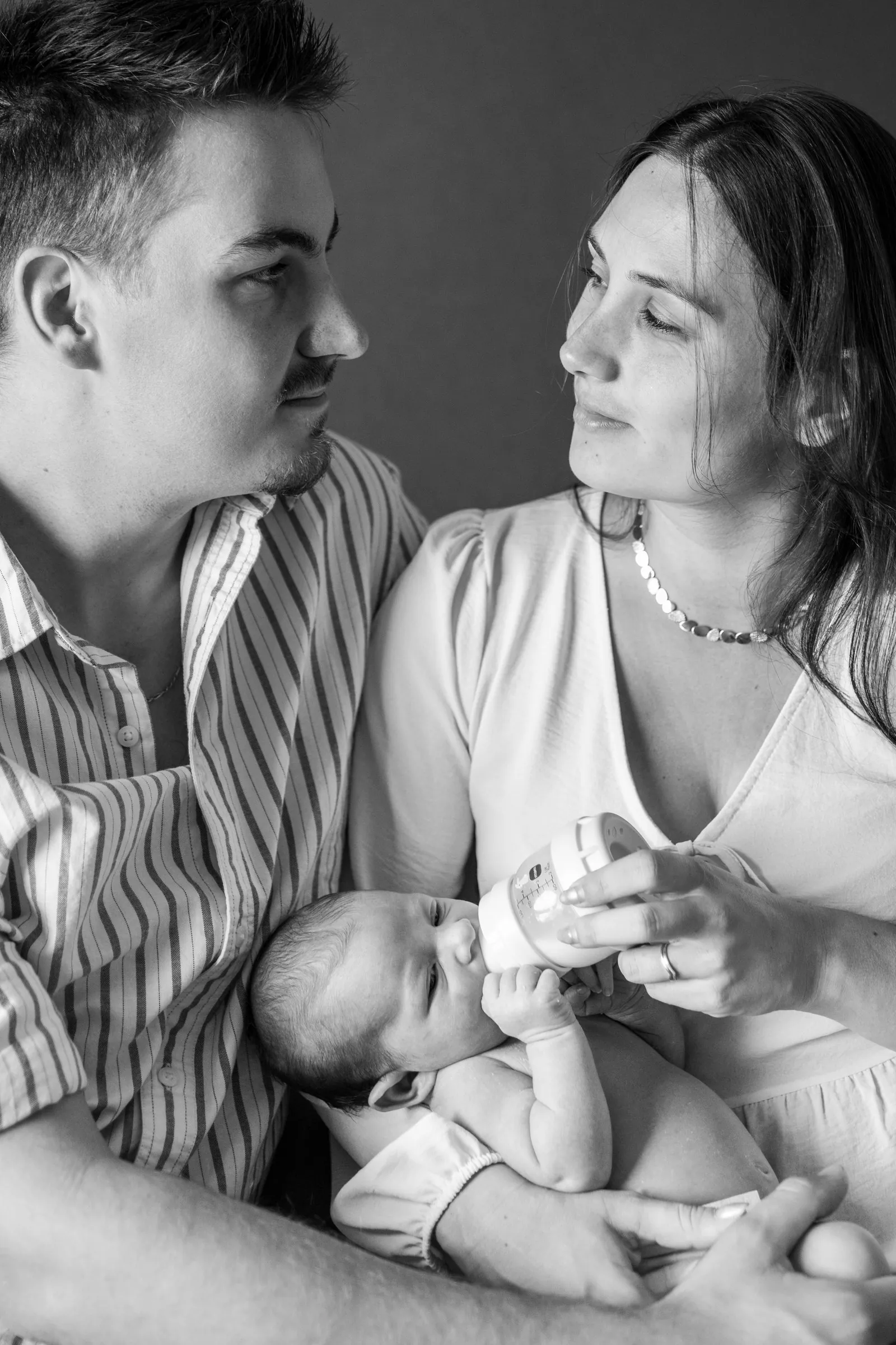 Séance naissance : parents qui échangent un regard pendant que maman donne le biberon au bébé, en noir et blanc