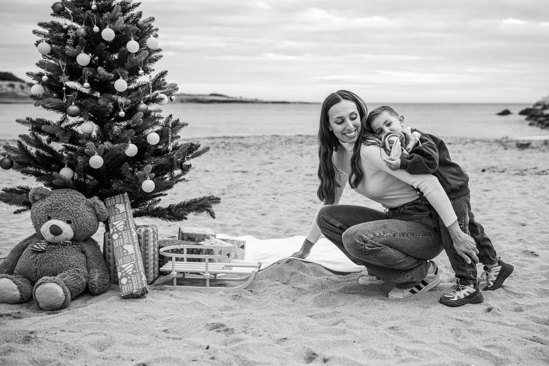 Christmas photo session on the beach: a mum and her son cuddling beside a decorated tree and presents, in black and white