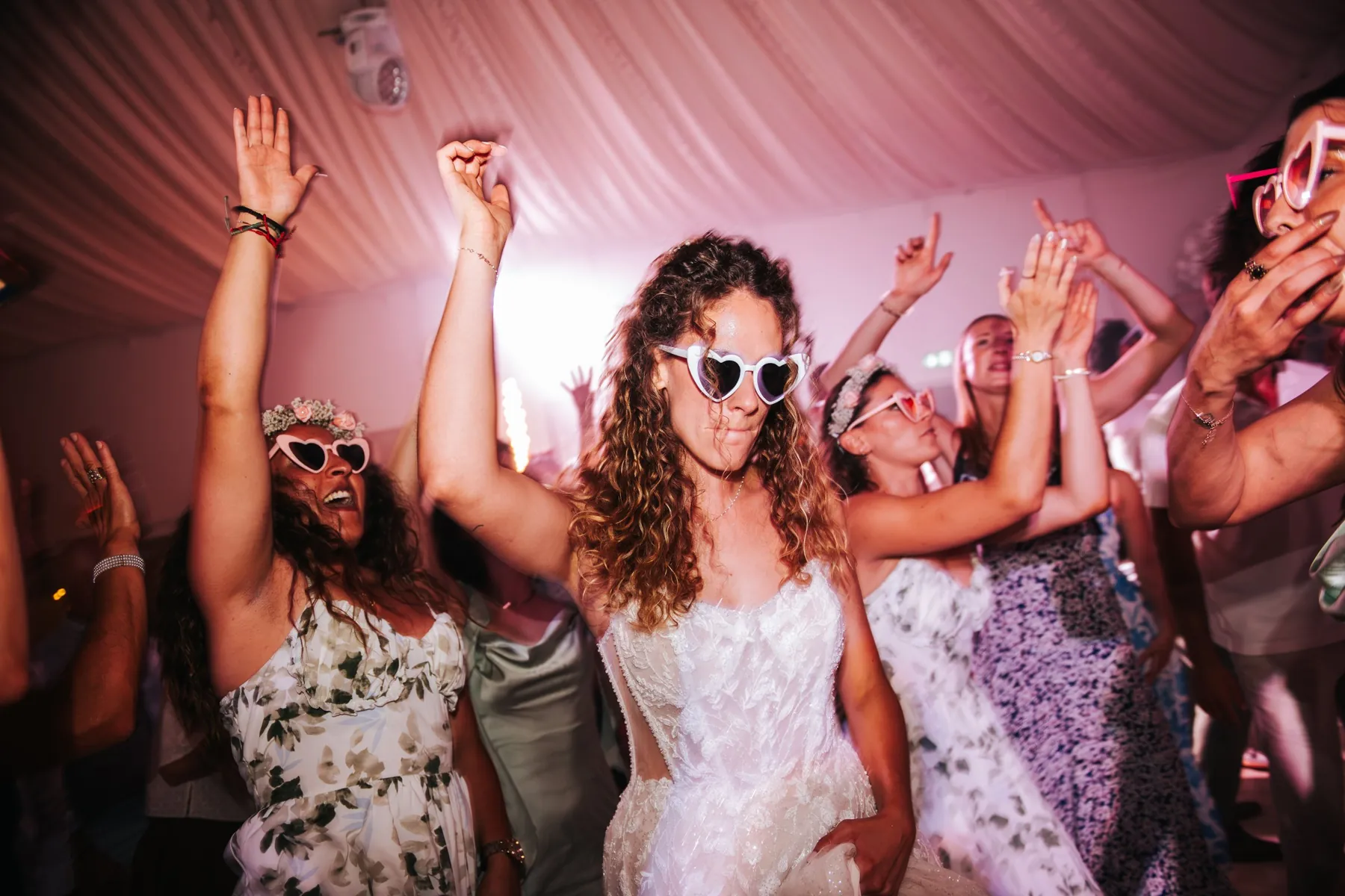 Soirée de mariage : la mariée danse avec ses amies, toutes en lunettes cœur, sous une lumière rose