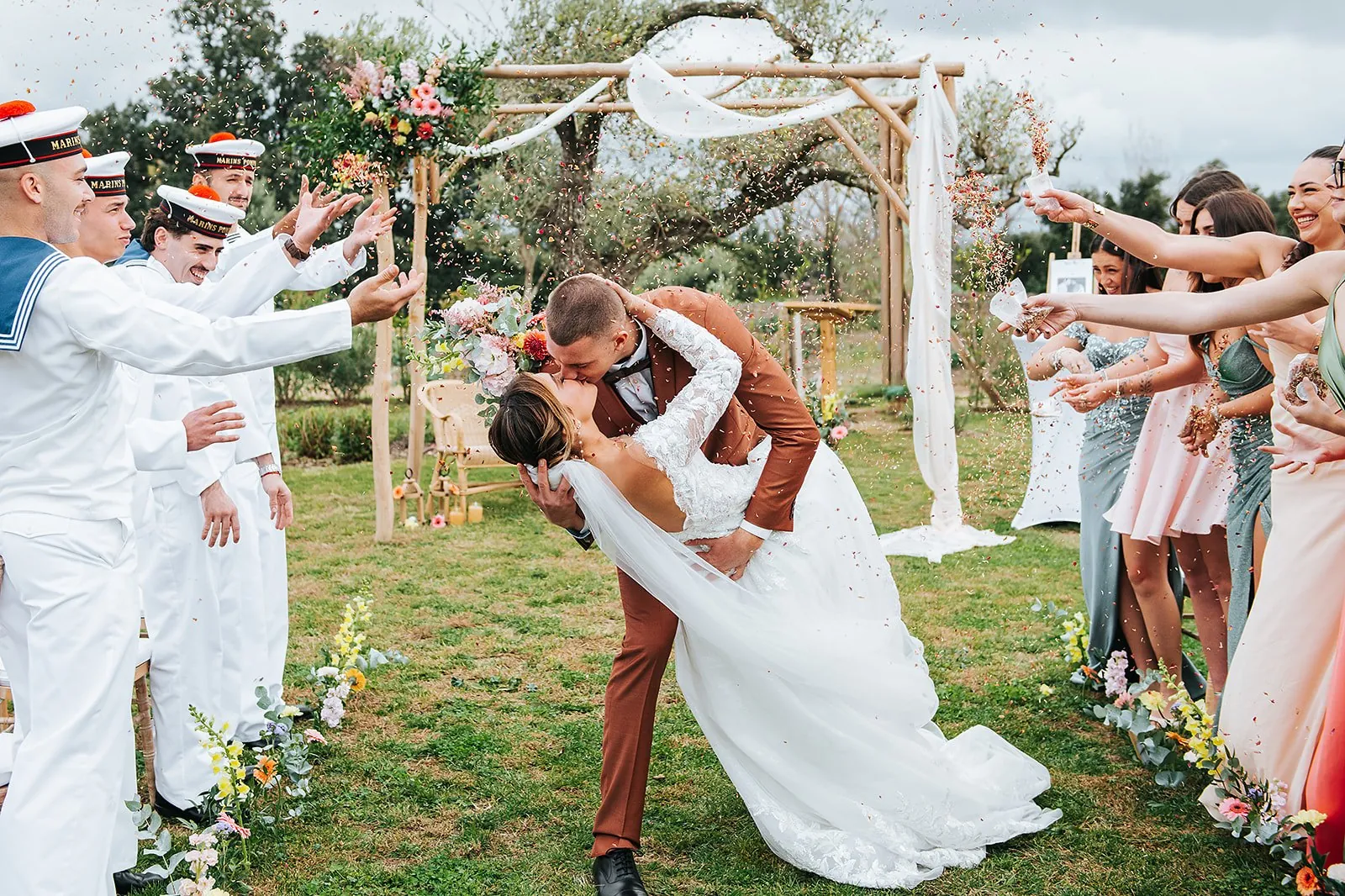 Sortie de cérémonie : le marié renverse la mariée pour un baiser sous une pluie de pétales lancés par les invités