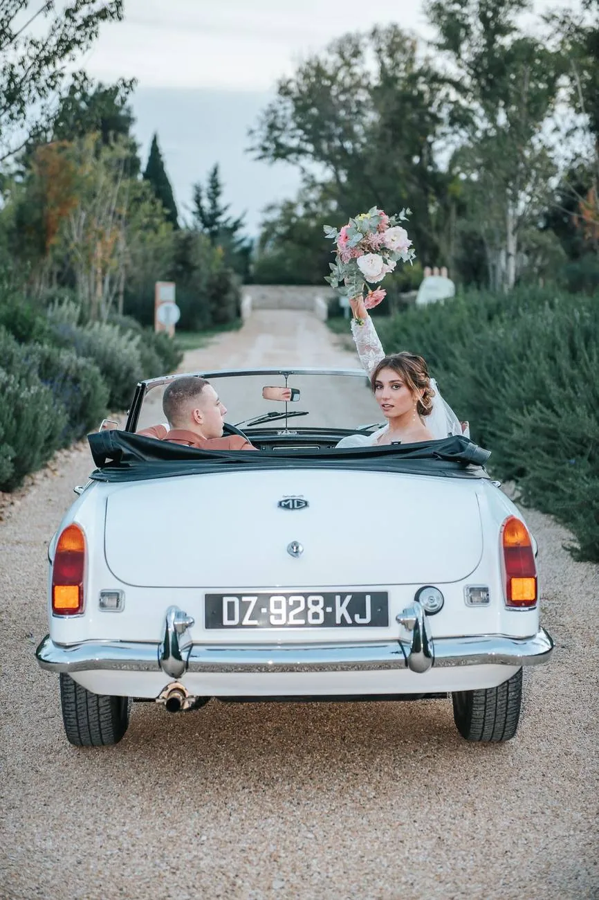 Sortie des mariés en cabriolet ancien blanc : la mariée lève son bouquet en se retournant sur une allée