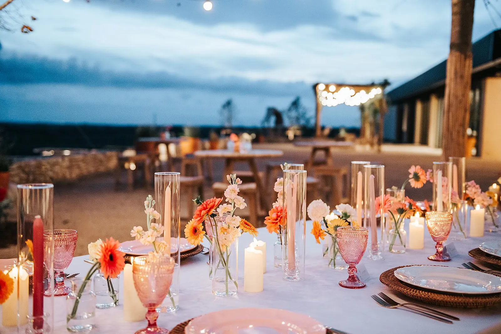 Table de réception de mariage à la tombée du jour : bougies, fleurs orangées, verres roses et guirlandes lumineuses