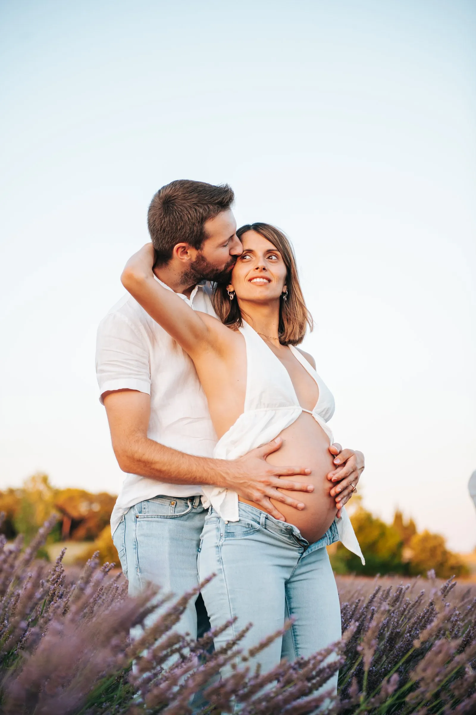 Maternity session in a lavender field: the father-to-be kisses the mother's temple, hands on her bump